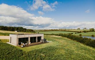 Modern wooden lodge with large glass windows and hot tub, set in a grassy hilltop overlooking green fields and a pond under a blue sky with scattered