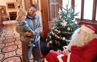 Father Christmas sitting beside a Christmas Tree, talking to a child being held by a woman.