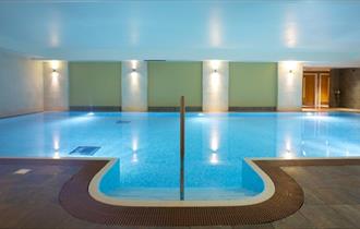 Large indoor swimming pool illuminated.
Dark tiled floor around the pool and 3 green walls at the back in-between sections with wall lights.
