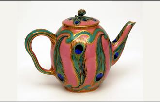 A colourful teapot with peacock feathers painted onto it.