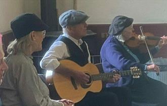 Folk musicians playing together in the Band Hall in The Pit Village.