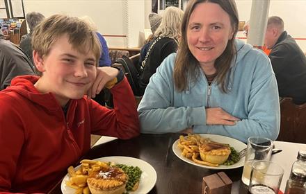 A woman and boy sitting at a table eating pie, chips and peas