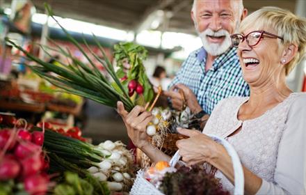 Woman and man smiling next to vegetable stall of market. Woman holding vegetables.