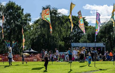 An audience view of the festival park with kids playing, a Bee Cart Walkabout artist, main stage, handmade Batik Flags.