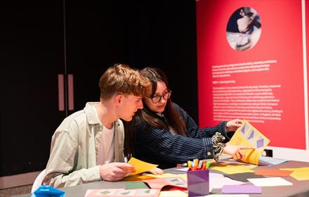 A young man with brown hair and a young woman with brown hair and glasses sat together making art in a bright pink space.