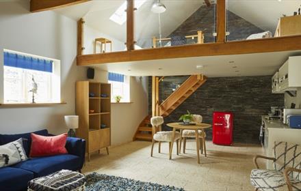 An open plan space with light flooring, dark blue sofa, small wooden table and 2 chairs. A kitchenette to the right with a red fridge.