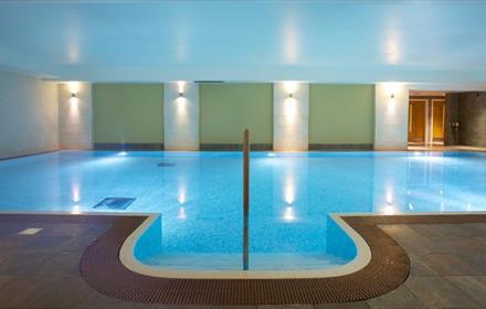 Large indoor swimming pool illuminated.
Dark tiled floor around the pool and 3 green walls at the back in-between sections with wall lights.