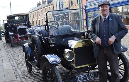 A man in period dress standing beside a vintage car in Beamish Museum's town