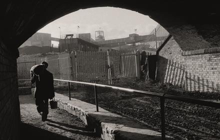 A photograph from the exhibition, a workman walking under railway bridge, carrying a bucket with a heavy sack over his shoulder