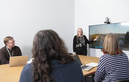 Meeting room with 4 people, three seated at table and one standing by display screen.