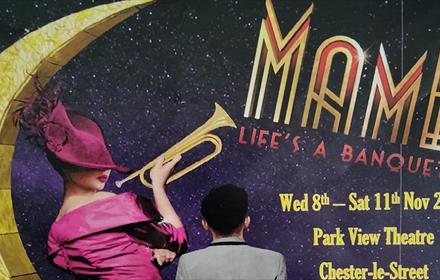 Image of a woman and a man sitting in front of a starry sky. The woman is wearing a fuchsia hat and dress and holding a trumpet.
