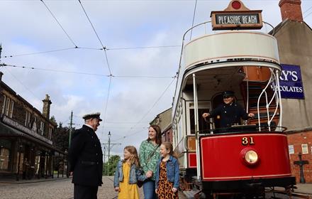 A mother and two children on Beamish 1900s Town Street with a tram and costumed Engagers.