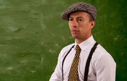 Man wearing a flat cap, white shirt, tie and braces.  Green background.