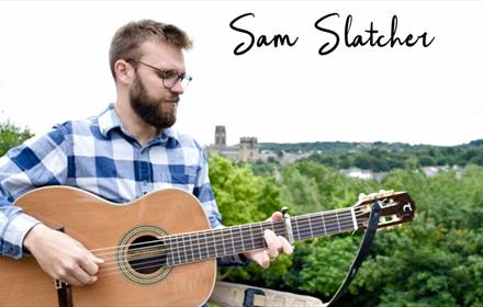 Sam Slatcher, musician, playing guitar with Durham Cathedral in the distance.