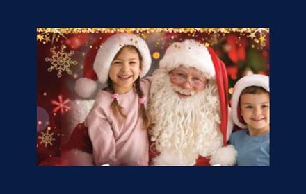 Young girl and boy on either side of Santa. Everyone is smiling. Girl and boy wearing Santa hats.