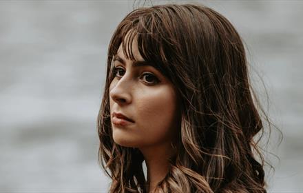 A women with brown hair looking out into the distance, there's a blurred grey background behind her.