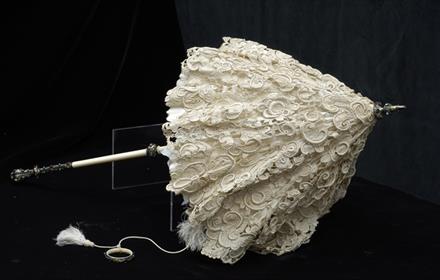An ornate 19th-century lace parasol with an intricately patterned cream lace canopy. The handle is made of ivory or bone with a decorative metal finia