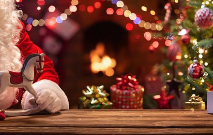 Santa holding a miniature rocking horse, and a Christmas tree, fairy lights and presents in the background.