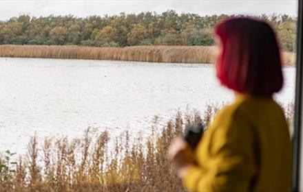 Woman with short red hair, wearing a yellow top, birdwatching with binoculars at RSPB Saltholme. Photo Credit: Brian Morrison (rspb-images.com)