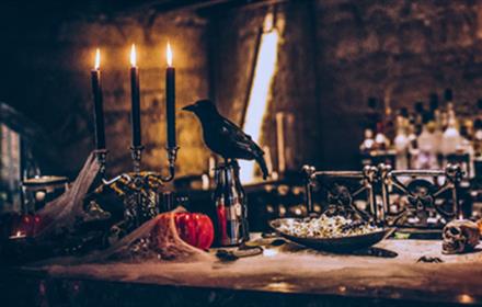 A raven sits at a dinner table covered in cobwebs, candles, skulls and pumpkins.
