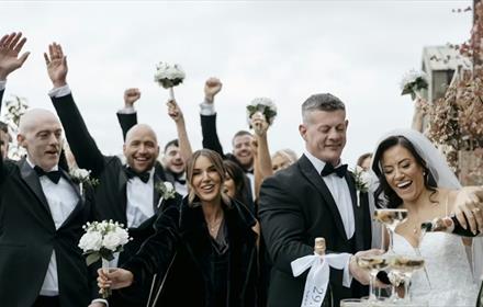 Bride and Groom smiling and pouring drinks, and their wedding party celebrating with their hands in the air behind them. Photo by Lauren Stacey.