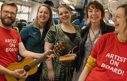 Sam Slatcher smiling and playing guitar on public transport, standing with four women who are also smiling.