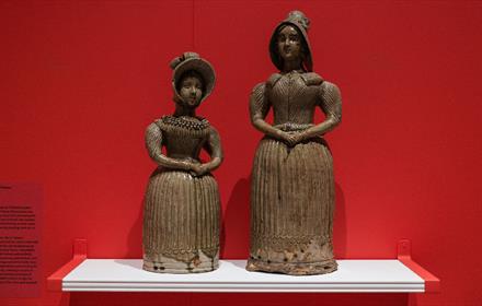 Two ceramic spirit flasks shaped like women in 19th-century attire stand on a white shelf against a vivid red wall, showcasing intricate detailing in