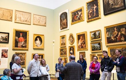 A group of ten people listening attentively to a curator in a museum gallery filled with framed historical paintings.