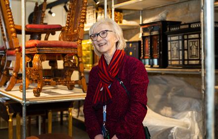 A smiling woman in a red outfit stands in a museum storeroom surrounded by intricately carved antique furniture and ornate wooden cabinets