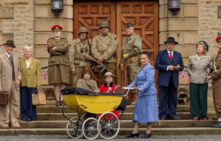 12 people in1940s clothing: men in suits and army uniform, women in skirts and trousers, and two children in coats and hats with a woman pushing a pra