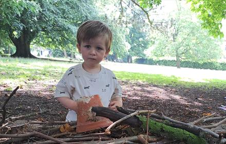 Child sitting in the grounds of The Bowes Museum , holding a plant pot,  surrounded by twigs and trees.