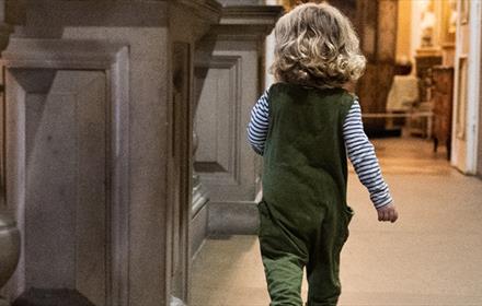 A young child with curly blonde hair, wearing green dungarees and a striped shirt, walks alone along a grand, ornately decorated museum hallway