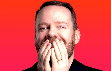 Headshot of Neil Delamere grinning with his hands covering his mouth.