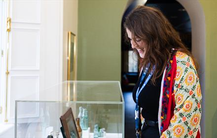 A woman wearing a colourful embroidered jacket smiles as she looks into a glass display case filled with vintage bottles and framed items in a museum