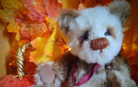 Handcrafted teddy bear sitting amongst autumn leaves and pumpkins.