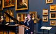 A woman standing at a lectern with a mic giving a speech in the picture gallery.