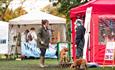 Two women stood talking with dogs on leads with market stalls in background.