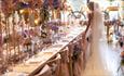 Image of a bride posing next to tables which have beautifully decorated with bouquets of flowers.