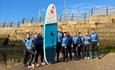 Group of paddleboarders in wetsuits posing with a paddleboard on a sandy beach near a stone wall under a clear blue sky.









