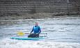 Smiling person kneeling on a paddleboard with a paddle, riding gentle waves near a stone sea wall.