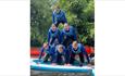 Group of young people in wetsuits forming a human pyramid on a paddleboard, smiling and having fun on the water.









