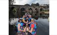 Group of people paddling a long boat on a calm river with an old stone bridge and riverside buildings in the background.









