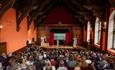 A full lecture hall with high wood-carved ceilings and a stage where two speakers present to an audience.