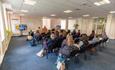 A bright presentation room with seated attendees facing a speaker at a podium and two large digital screens.
