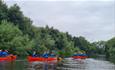 4 red canoes in the river, filled with participants wearing blue life jackets.
The river is lined with green trees.