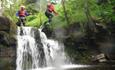 2 children in life vests and safety helmets jumping from some rocks into a river with a small waterfall below them