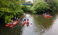 Group of people in red canoes paddling along a tree-lined river with a blue footbridge in the background.