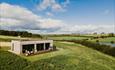Modern wooden lodge with large glass windows and hot tub, set in a grassy hilltop overlooking green fields and a pond under a blue sky with scattered