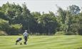 A golfer pulling his golf bag on a golf trolley across the green. Large trees are in the distance.