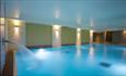 A side angle of the indoor swimming pool that is illuminated.
A fountain of water sprays over the side and the walls are lit with wall lights.